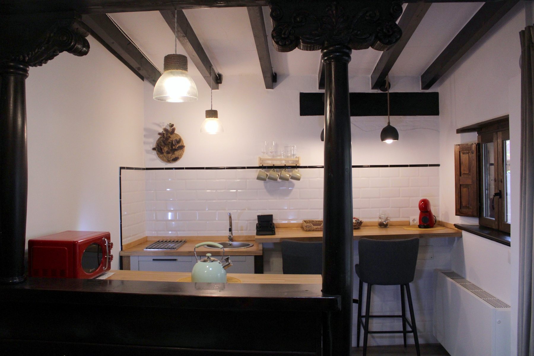 Cocina con azulejos blancos, microondas rojo, tetera y barra con taburetes