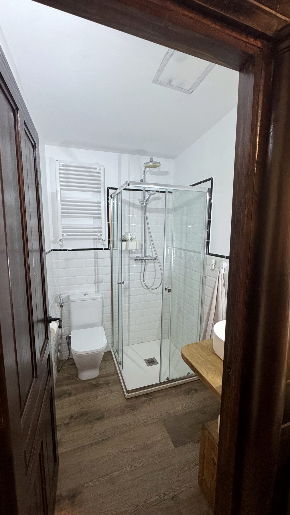 Baño con ducha de mampara, azulejos blancos y suelo de madera